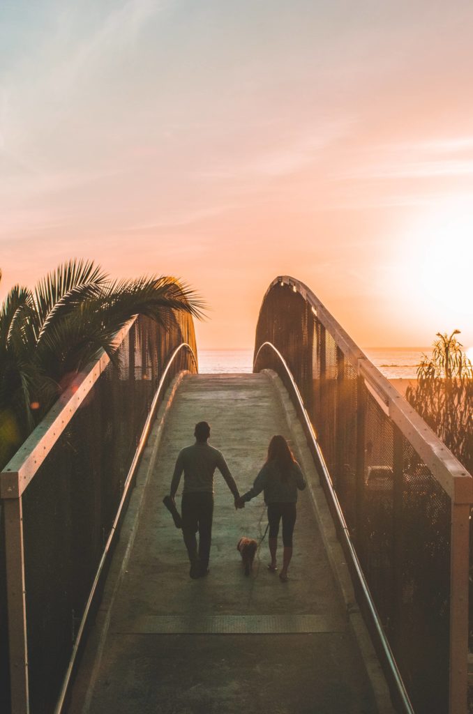 couple promenant un chien, traversant un pont face à la mer, au soleil couchant